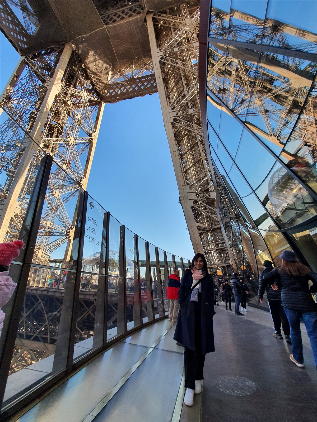 A woman in a long dark coat walking along the glass-walled gallery of the Eiffel Tower's first level