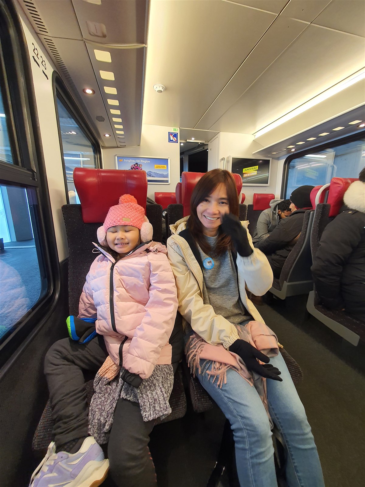 A girl in a pink hat and a woman in a cream jacket sitting in red-seated Jungfrau Railway carriage