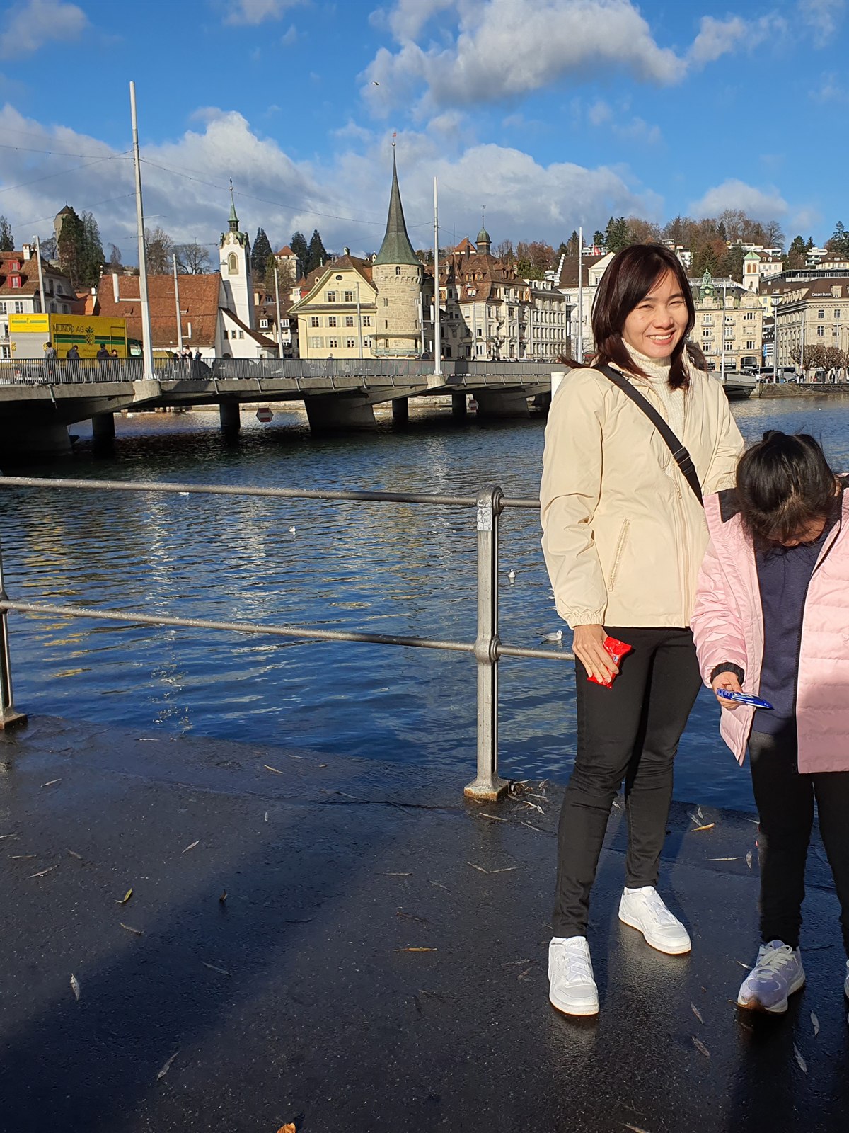 Two girls standing on the lakeside promenade in Lucerne, with a bridge and old town buildings across the water