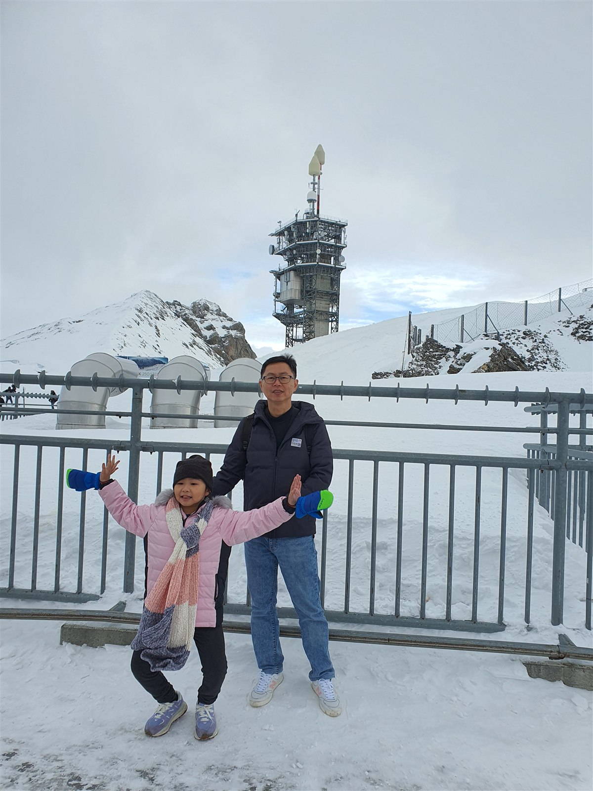 Father and daughter with arms out in front of the summit railing and the mountain transmission tower