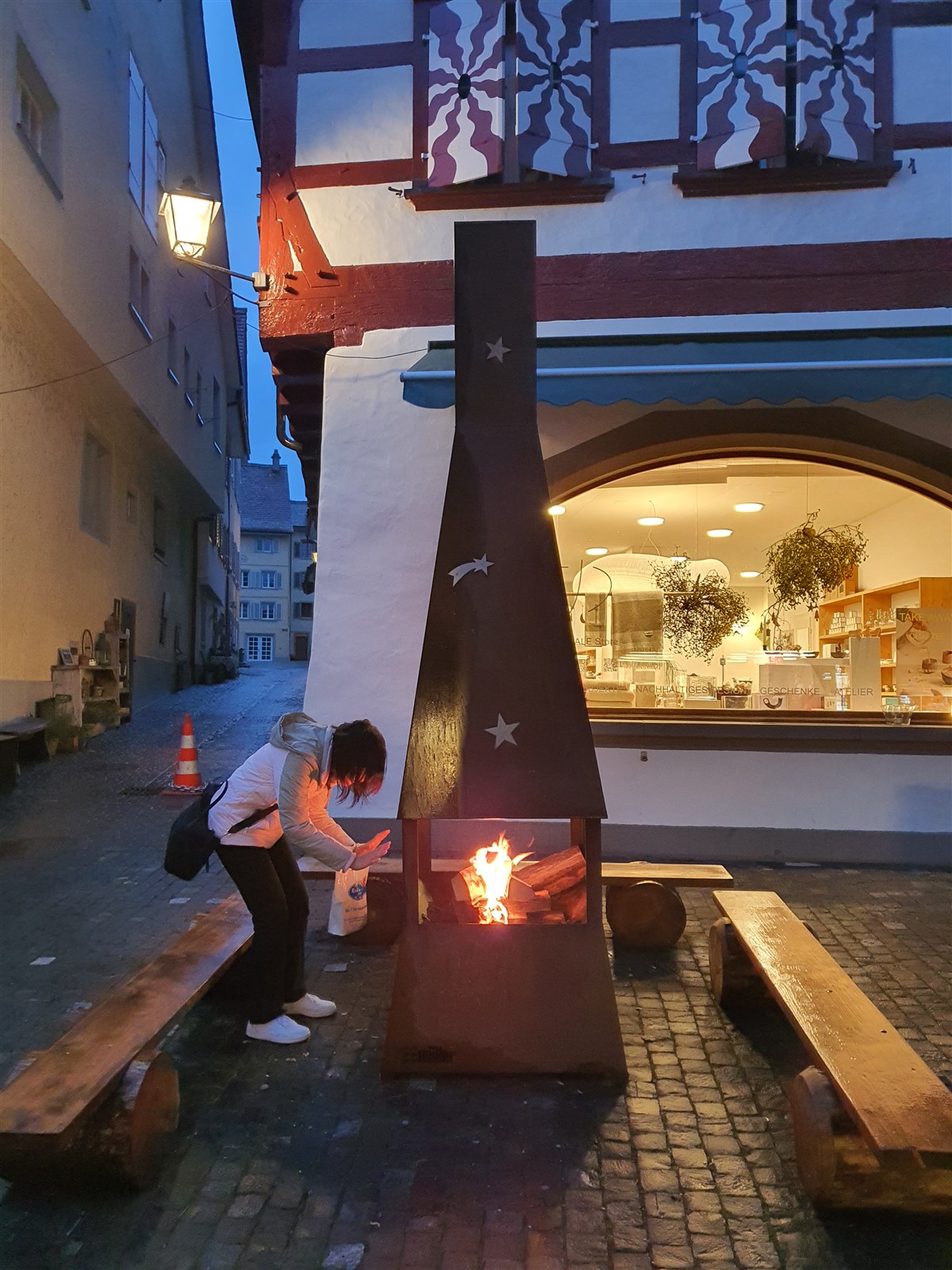 A woman warming her hands at an open fire on a bench in Stein am Rhein at dusk