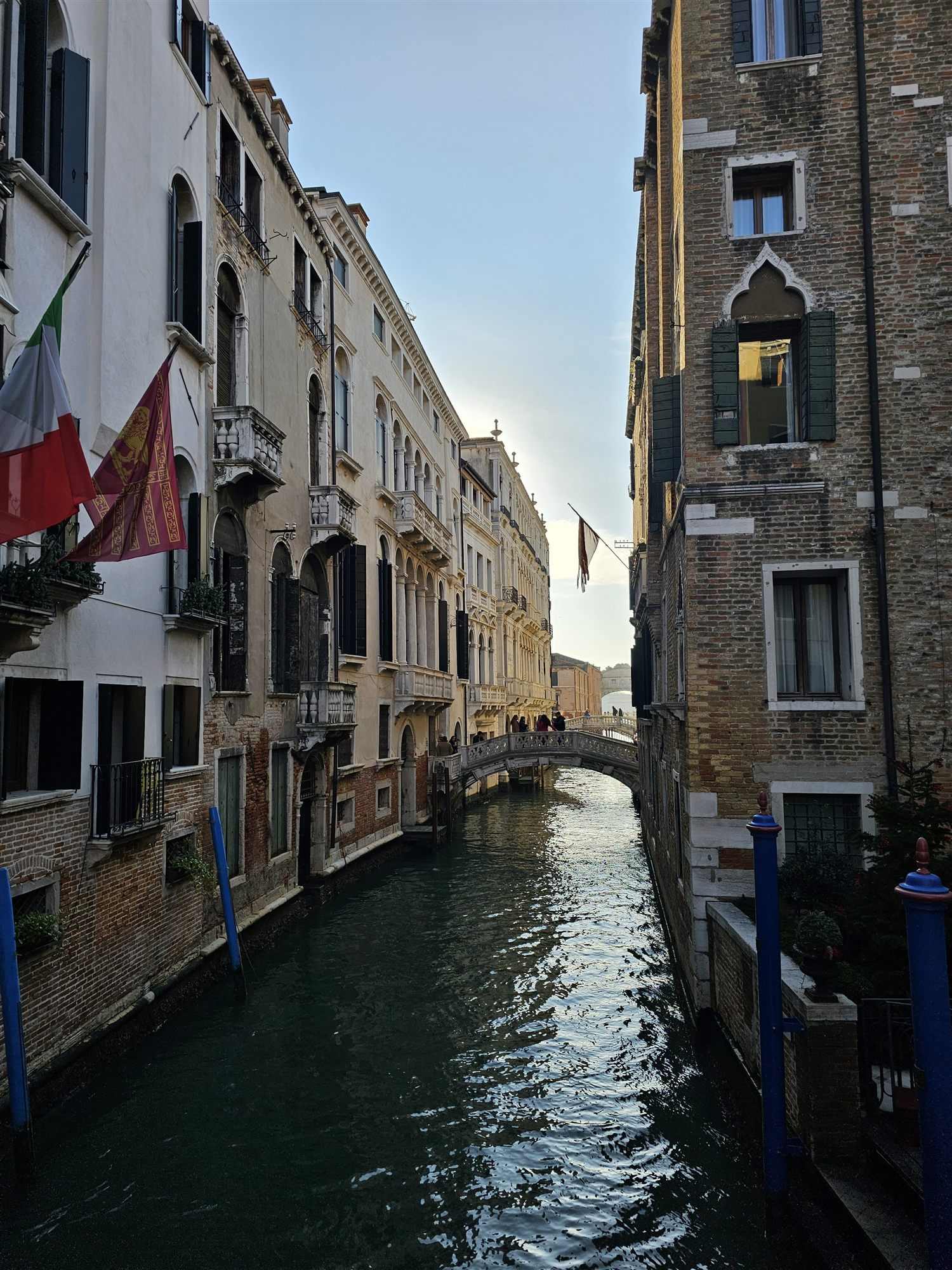 A narrow Venice canal with flags on the palazzo walls and the Bridge of Sighs in the distance